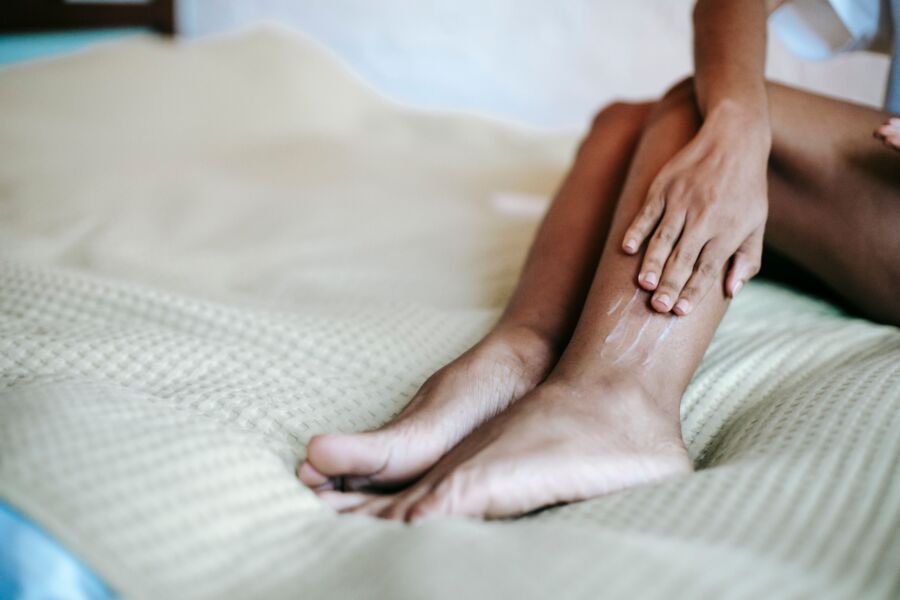 Recorte crema untable femenina irreconocible en las piernas mientras está sentado en la cama en casa