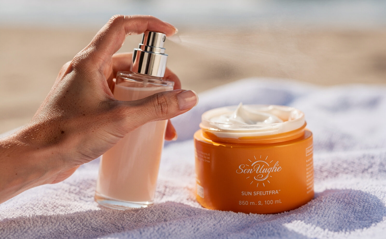 Primer plano de una mano pulverizando un frasco de perfume sin etiqueta ni marca visible (el gesto y el envase desenfocado o de espaldas a cámara), junto a un bote de crema solar abierto sobre una toalla de playa, fondo de arena desenfocada, luz de mediodía. El foco visual está en el contraste frasco de perfume/bote de crema solar, no en identificar el producto. Sin segunda persona, sin frasco flotando sobre fondo blanco, sin marca ni etiqueta legible en ningún envase.