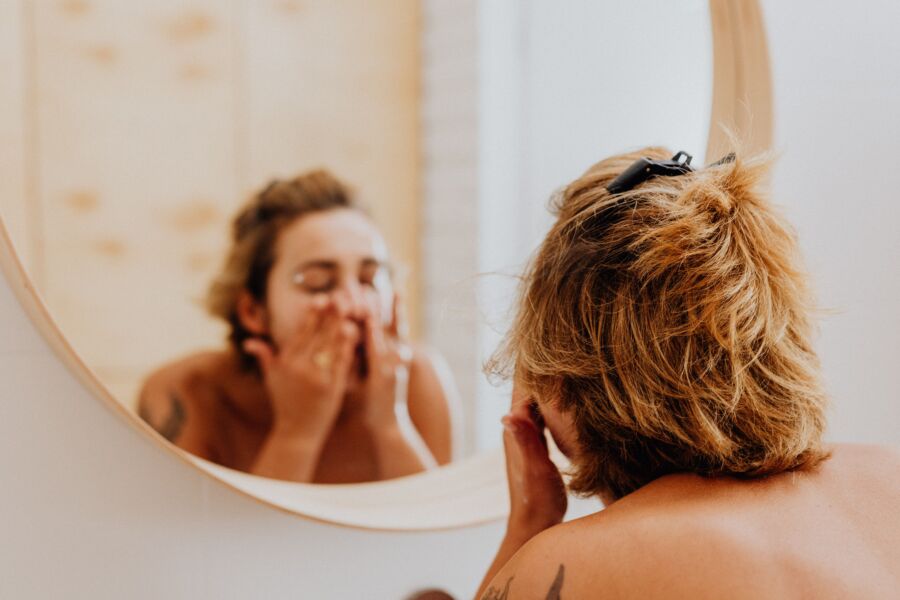 Mujer joven en el baño aplicando productos faciales mientras se mira en el espejo, abrazando el autocuidado.