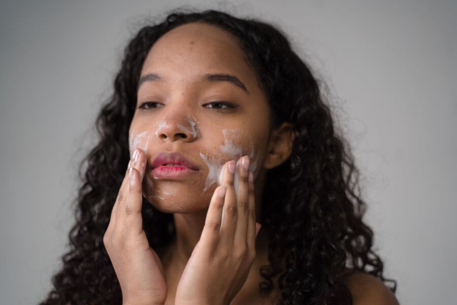 Mujer afroamericana de pelo largo oscuro rizado lavando mejillas con espuma facial sobre fondo gris