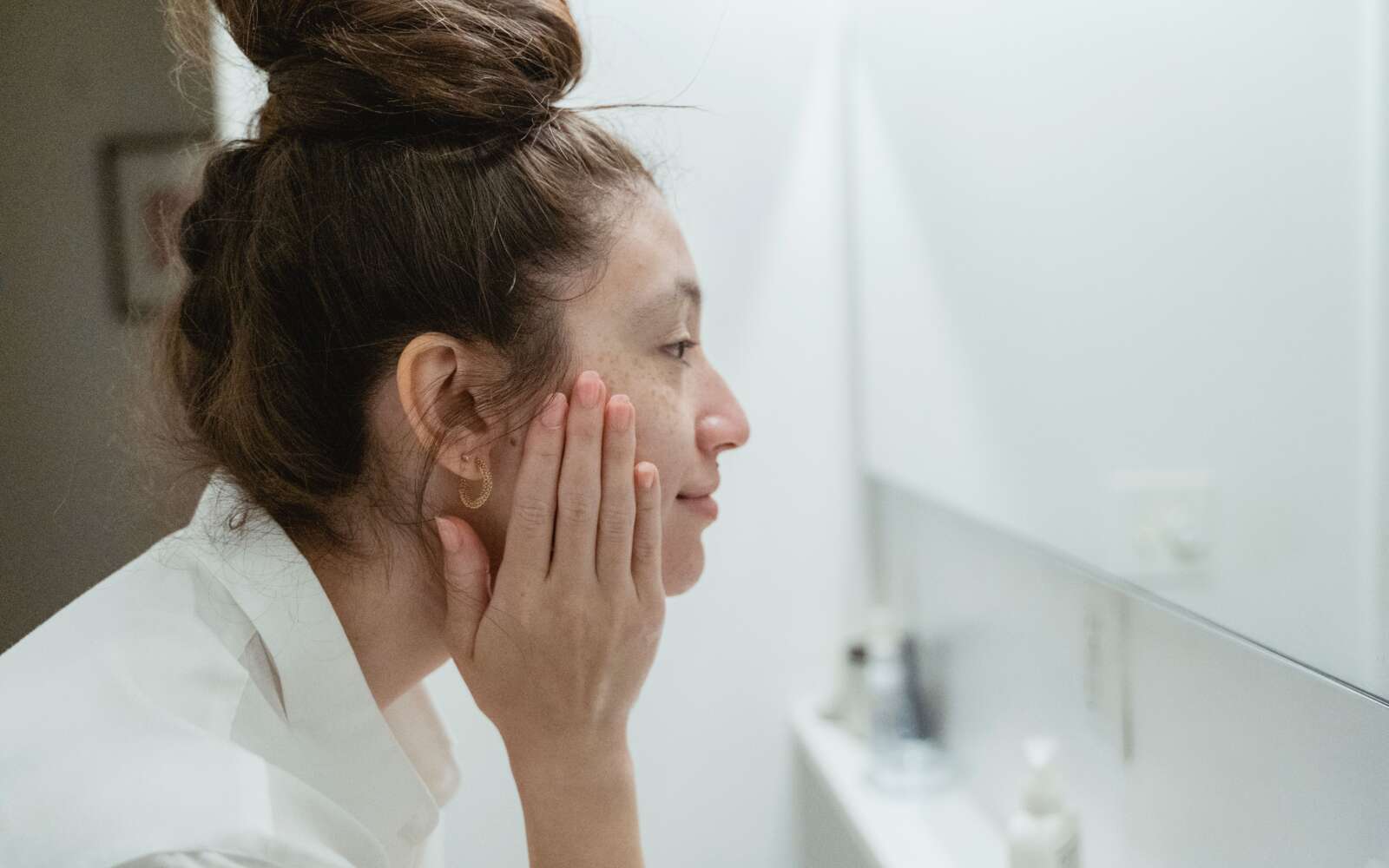 Vista de perfil de una mujer tocándose la cara y reflejándose en el espejo del baño.
