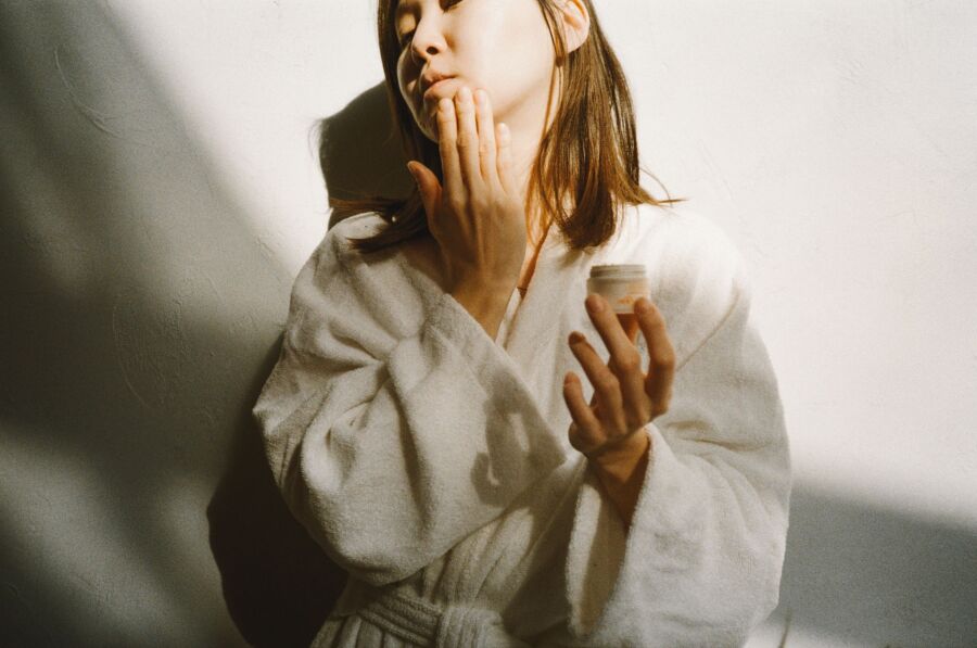 Mujer asiática en bata blanca aplicando crema facial en un ambiente interior iluminado.