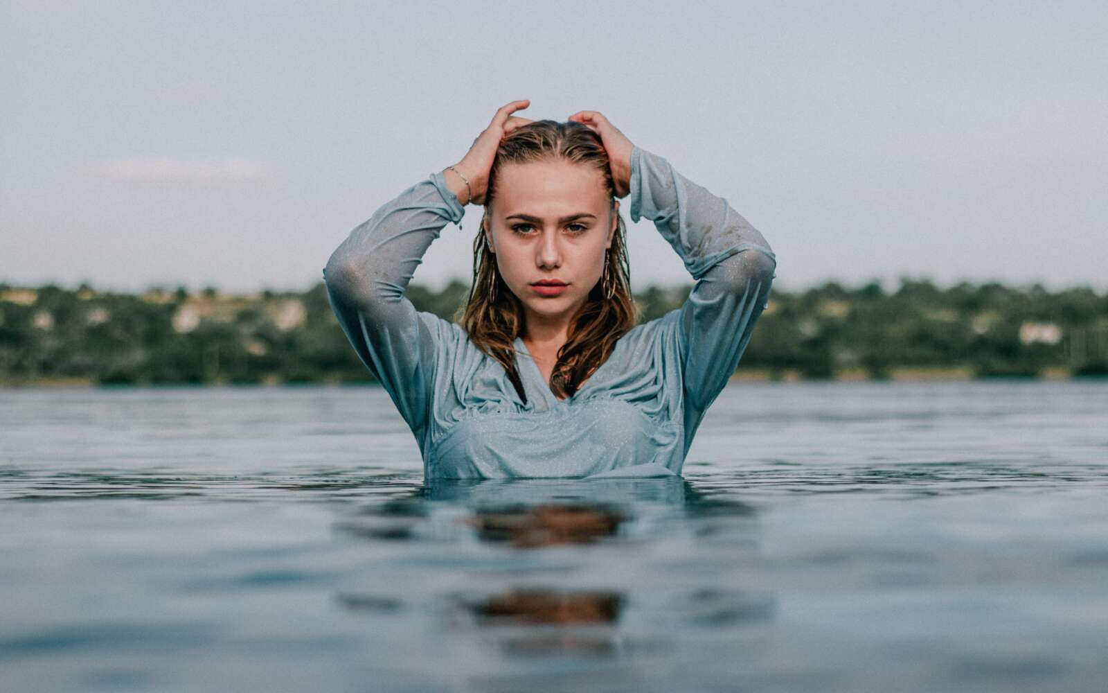 Una mujer de pie en aguas tranquilas con las manos en el pelo y expresión serena.