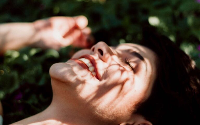 Primer plano de una mujer sonriente disfrutando de la luz del sol, con sombras que crean una sensación serena al aire libre.